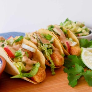 Plate of three fish stick tacos with slaw and aioli, fresh cilantro garnish, beside lime halves, inviting, easy family dinner ready to devour.