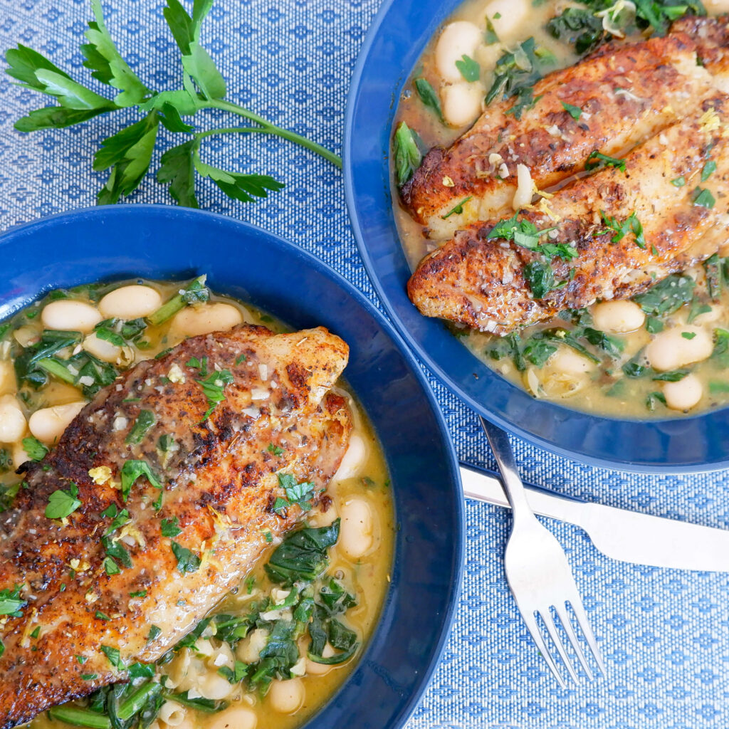 Seared white fish fillet on top of smoky butter beans and wilted spinach in a shallow bowl, drizzled with pan sauce and lemon zest